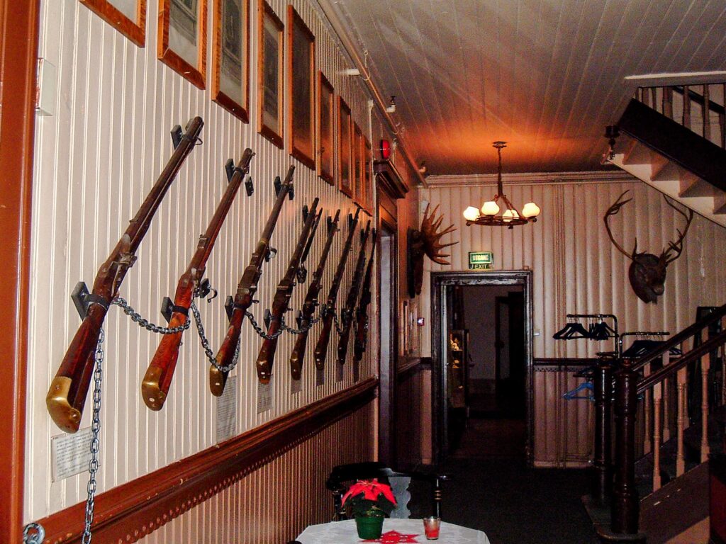 Interior de um hall de hotel, com uma escadaria à direita, uma parede enfeitada com rifles à esquerda e duas cabeças de alces empalhadas nas paredes.